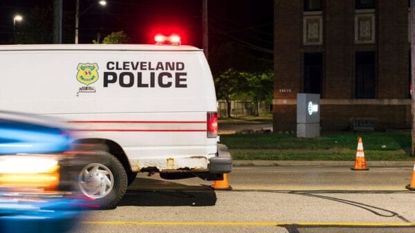 A Cleveland Police van is parked on a city street at night, with orange traffic cones nearby and a blurred blue car passing in the foreground.