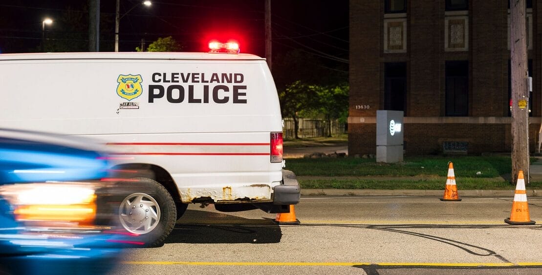 A Cleveland Police van is parked on a city street at night, with orange traffic cones nearby and a blurred blue car passing in the foreground.