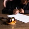 A judge sits at a desk writing on papers, with a wooden gavel resting on a sound block in the foreground.