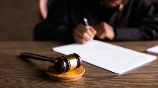 A judge sits at a desk writing on papers, with a wooden gavel resting on a sound block in the foreground.