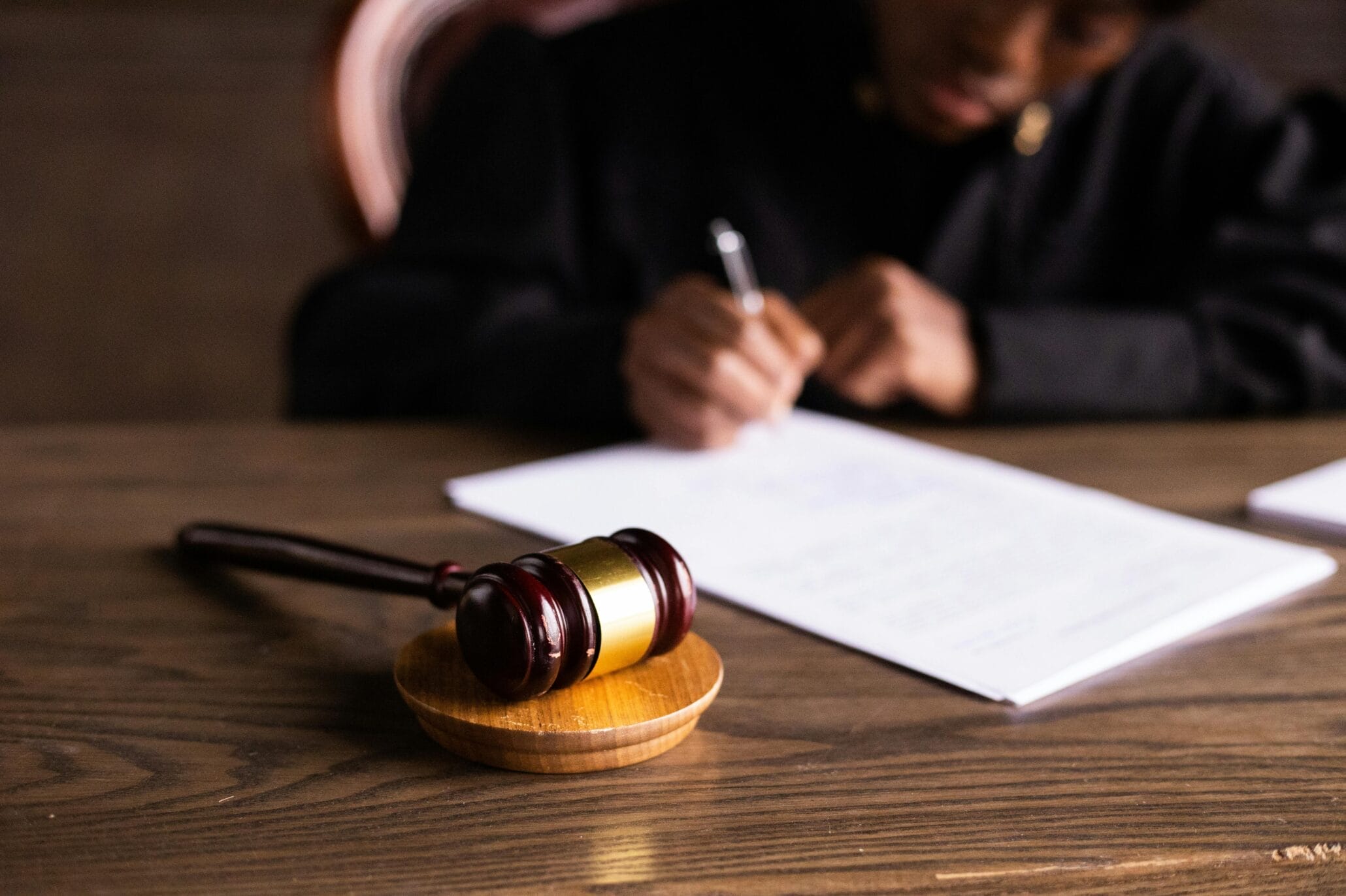 A judge sits at a desk writing on papers, with a wooden gavel resting on a sound block in the foreground.