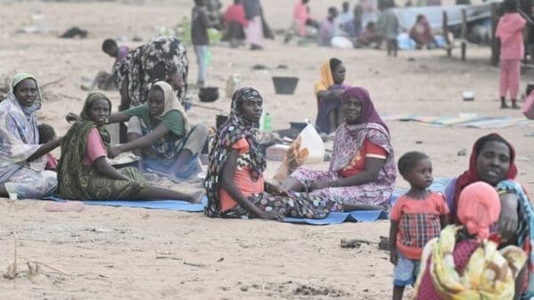 Groups of women and children sit and interact on sandy ground outdoors, some on mats, with sparse belongings and tents visible in the background.