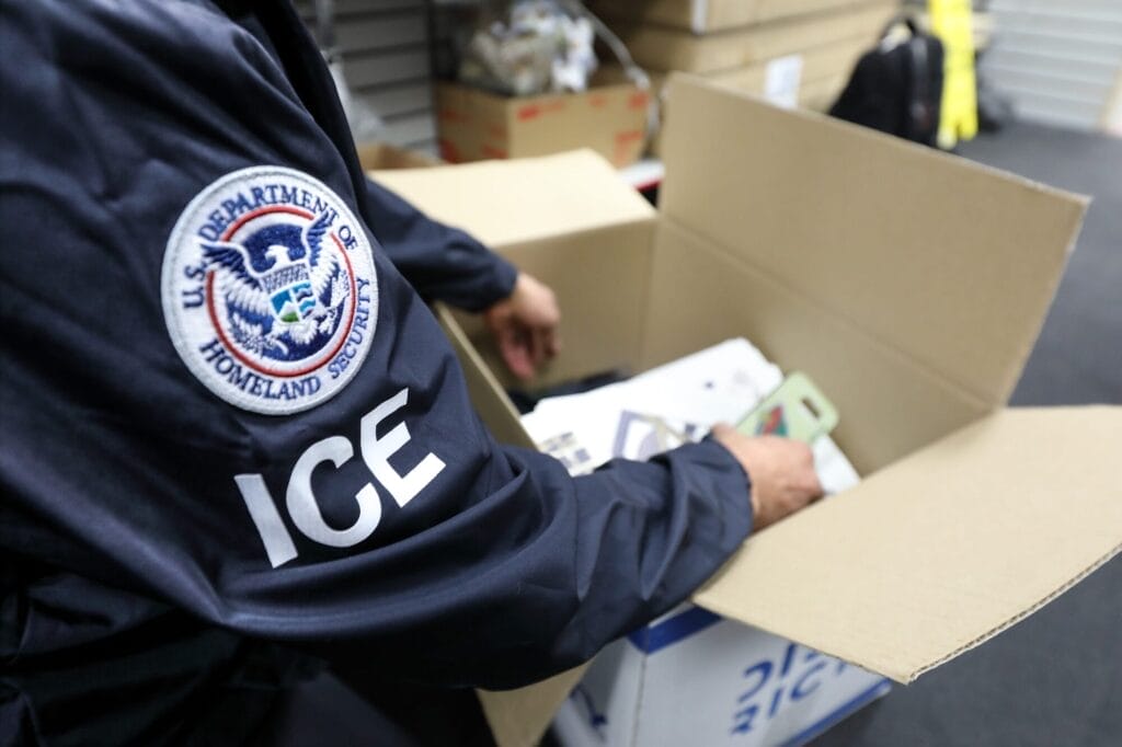 A person wearing an ICE jacket with a Homeland Security patch inspects items inside an open cardboard box in an indoor setting.