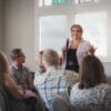 A woman speaking into a microphone stands in front of a group of seated people in a bright room, engaging with her audience.